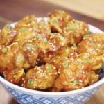 Bowl filled with crispy honey butter fried chicken pieces coated in a sticky glaze and topped with sesame seeds and scallions.