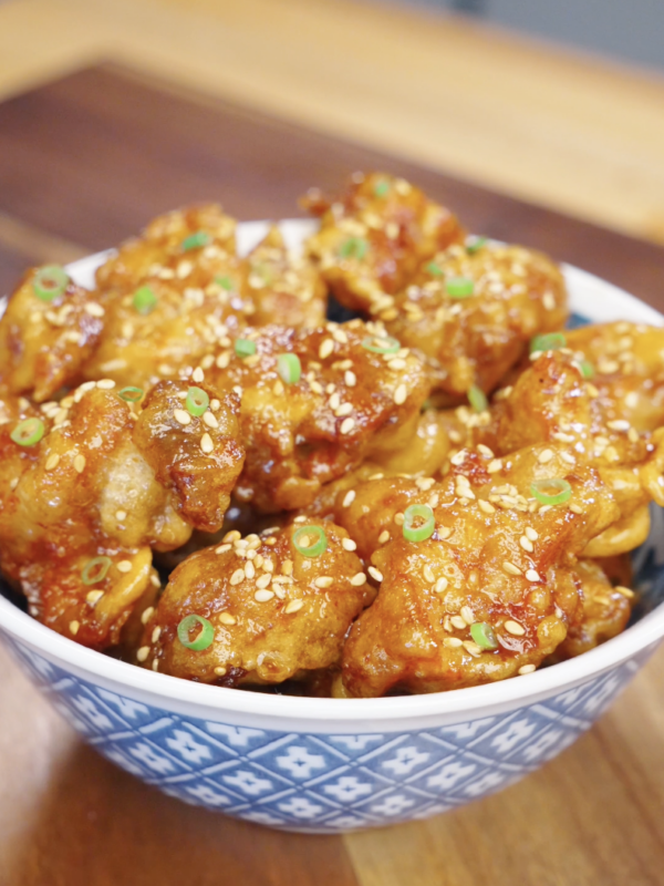 Bowl filled with crispy honey butter fried chicken pieces coated in a sticky glaze and topped with sesame seeds and scallions.