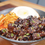 Bowl of Korean BBQ steak over rice topped with sesame seeds, scallions, kimchi, and a fried egg, highlighting glossy caramelized beef.