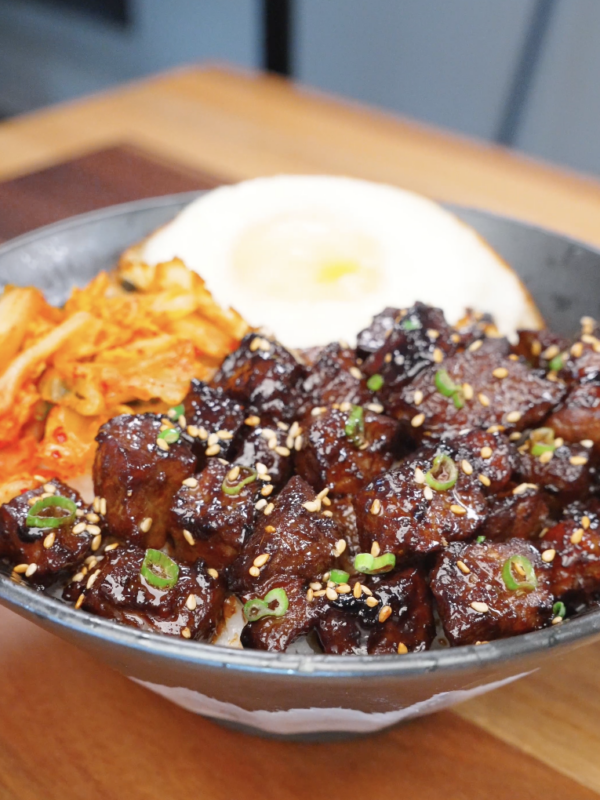 Bowl of Korean BBQ steak over rice topped with sesame seeds, scallions, kimchi, and a fried egg, highlighting glossy caramelized beef.