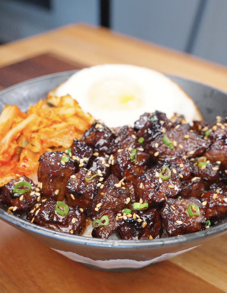 Bowl of Korean BBQ steak over rice topped with sesame seeds, scallions, kimchi, and a fried egg, highlighting glossy caramelized beef.