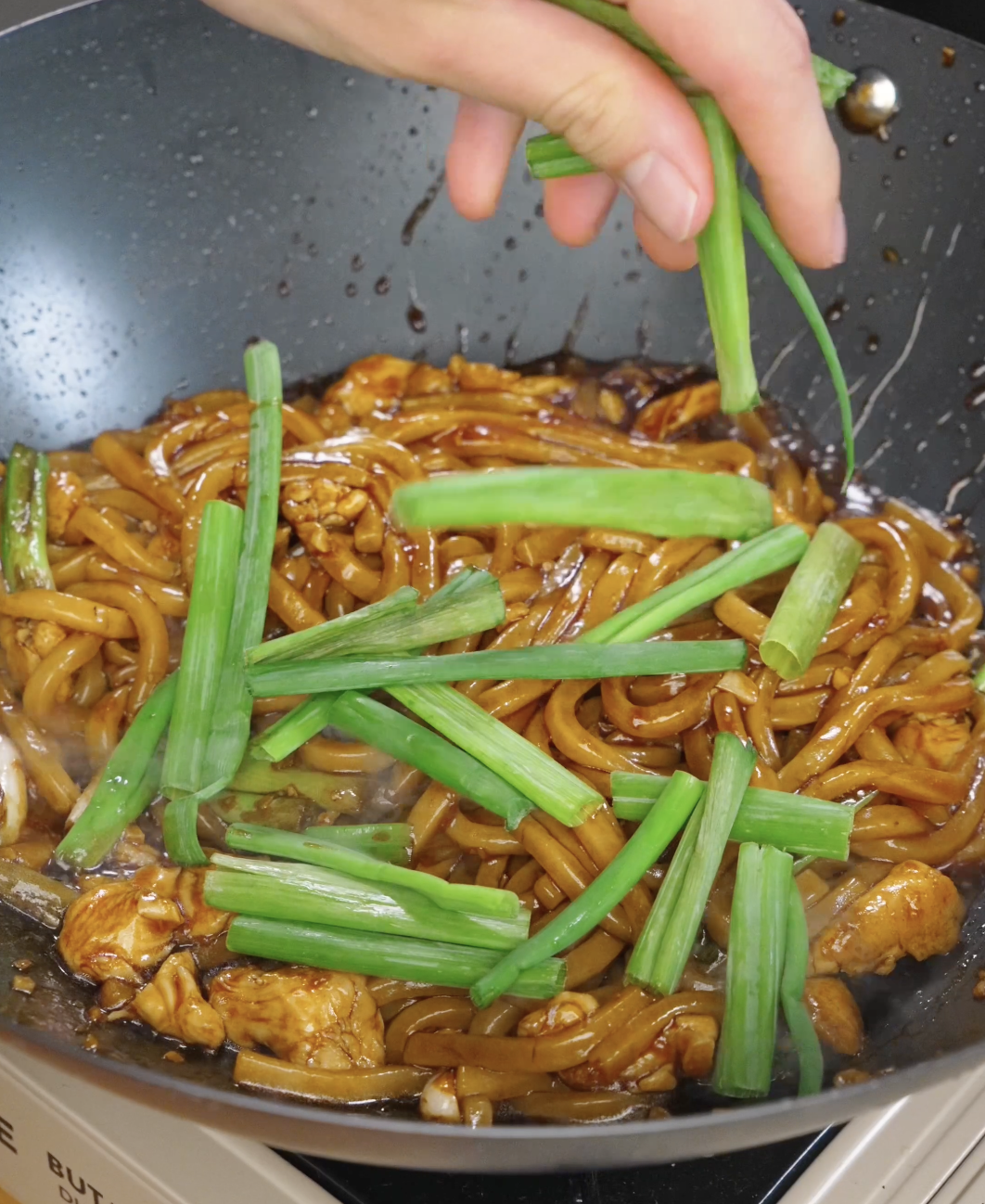 Fresh scallions being added to garlic chicken noodles in a pan before serving.
