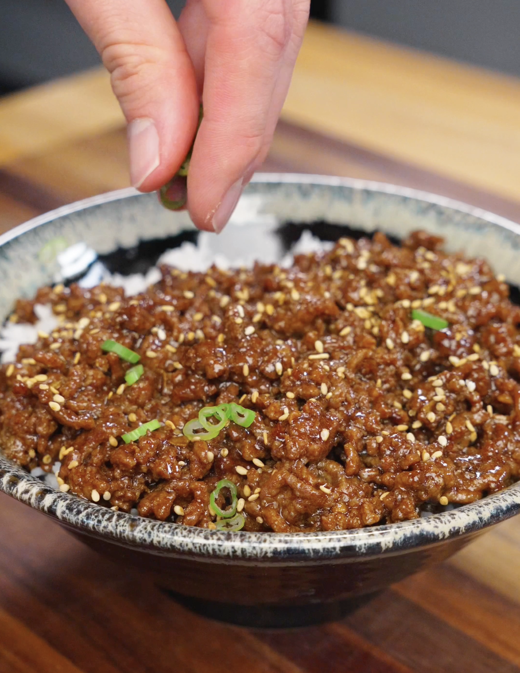 Hand sprinkling sliced scallions over a bowl of bulgogi ground beef with sesame seeds.