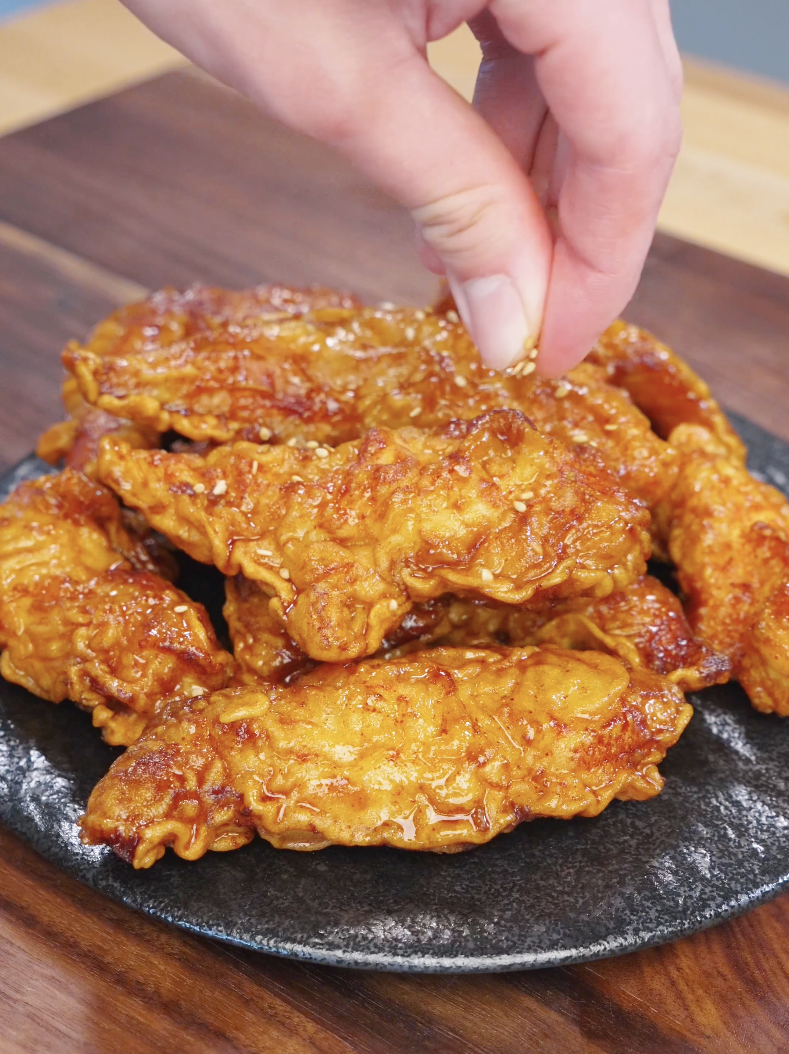 Hand sprinkling sesame seeds over freshly glazed honey butter chicken tenders on a plate.