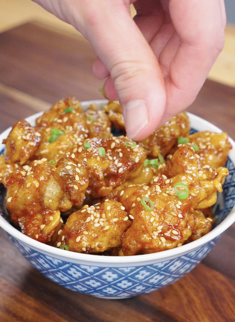 Hand sprinkling sesame seeds and chopped scallions over freshly glazed honey butter fried chicken in a bowl.