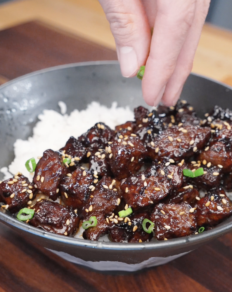 Sprinkling sesame seeds and chopped scallions over Korean BBQ steak on rice, adding texture and finishing the dish with fresh garnish.