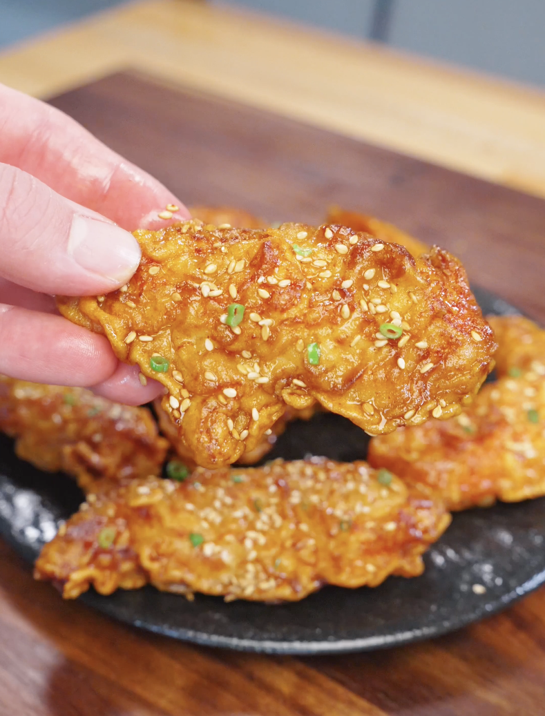 Hand holding a golden crispy honey butter chicken tender coated in sticky glaze with sesame seeds.