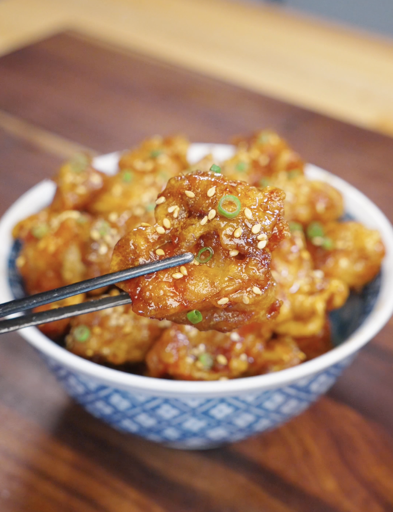 Chopsticks holding a crispy piece of honey butter fried chicken coated in a glossy sweet sauce with sesame seeds and scallions.