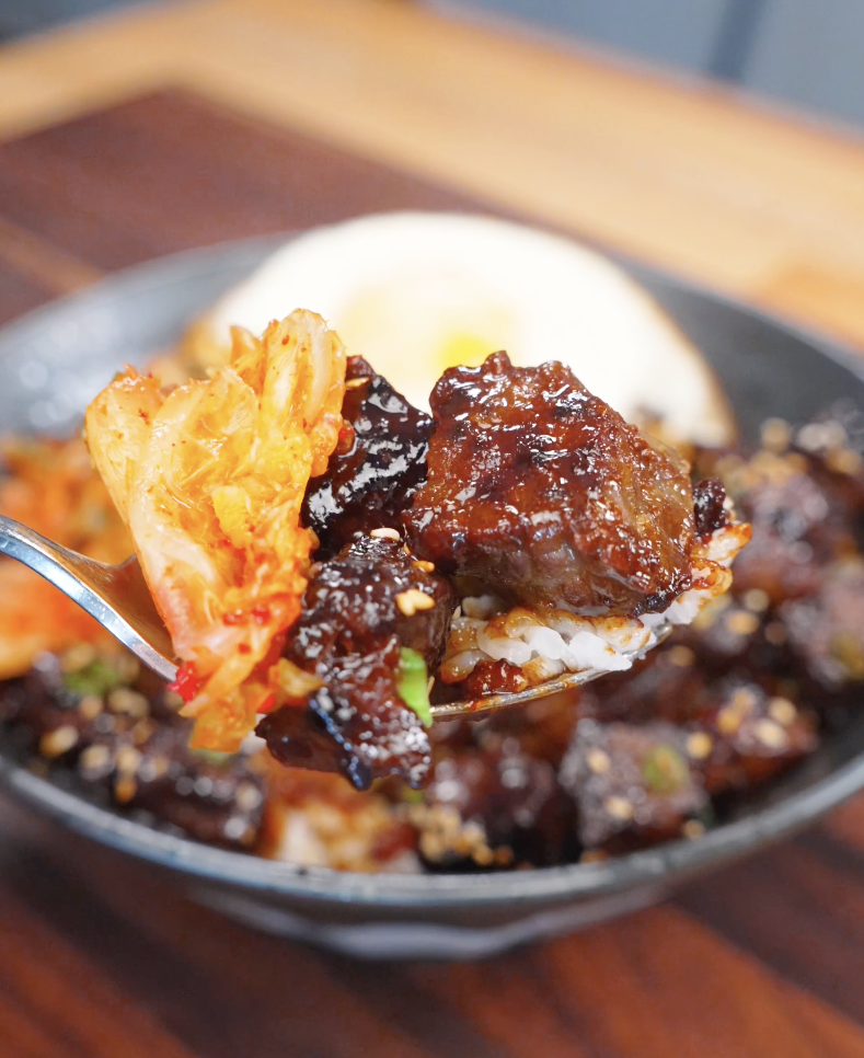 Spoonful of Korean BBQ steak served over white rice with kimchi, showing tender glazed beef and vibrant contrast of textures and flavors.