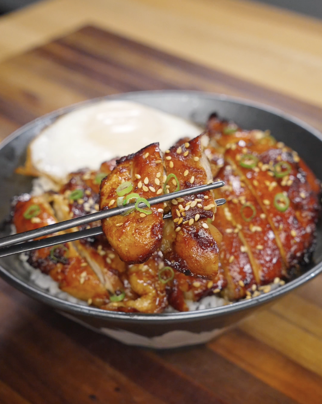 Chopsticks lifting a piece of caramelized chicken bulgogi coated in a glossy gochujang-based sauce, topped with sesame seeds and sliced scallions over a bowl of rice.