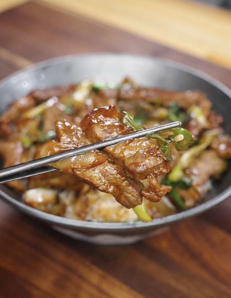 Chopsticks holding a glossy slice of Mongolian beef over rice, showing caramelized sauce coating with scallions and onions in this Mongolian Beef Recipe.