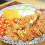 Bowl of steamed rice topped with crispy pork belly slices and arranged with fresh cucumbers, cherry tomatoes, and pickled vegetables in this Vietnamese Crispy Pork Belly Recipe.