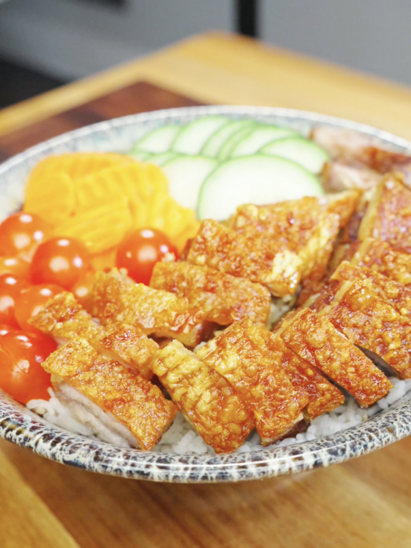 Bowl of steamed rice topped with crispy pork belly slices and arranged with fresh cucumbers, cherry tomatoes, and pickled vegetables in this Vietnamese Crispy Pork Belly Recipe.