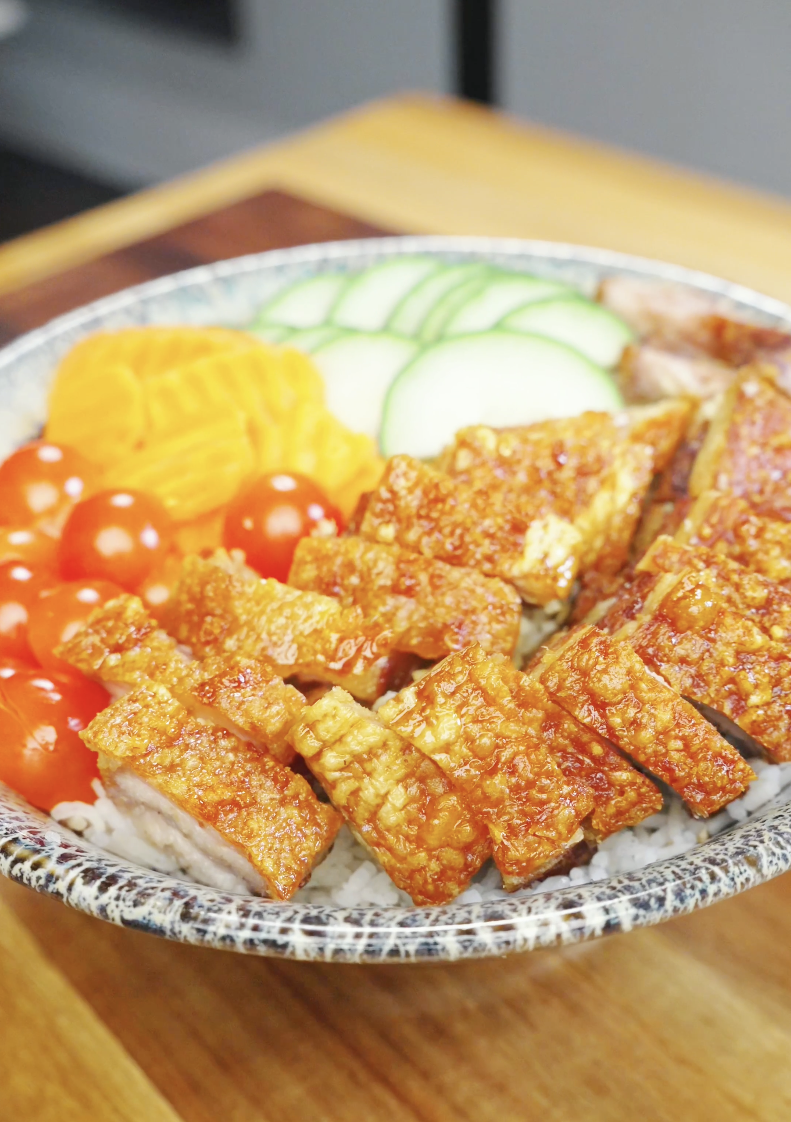 Bowl of steamed rice topped with crispy pork belly slices and arranged with fresh cucumbers, cherry tomatoes, and pickled vegetables in this Vietnamese Crispy Pork Belly Recipe.