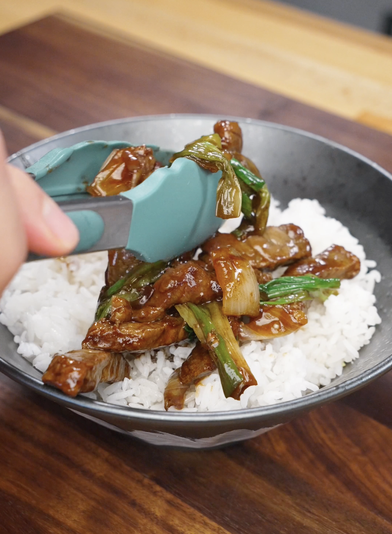 Glossy Mongolian beef being placed over steamed white rice in a bowl, with onions and scallions completing the Mongolian Beef Recipe.