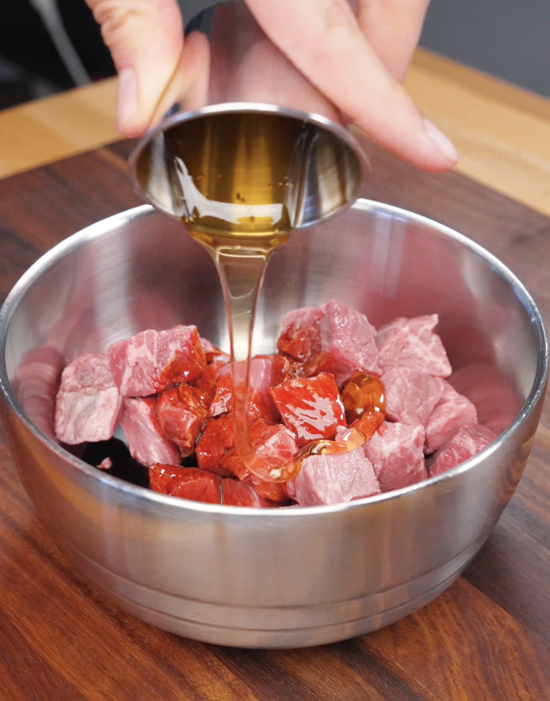 Honey being poured over cubed steak in a bowl, coating the beef with soy sauce, honey, and aromatics for Korean BBQ flavor.