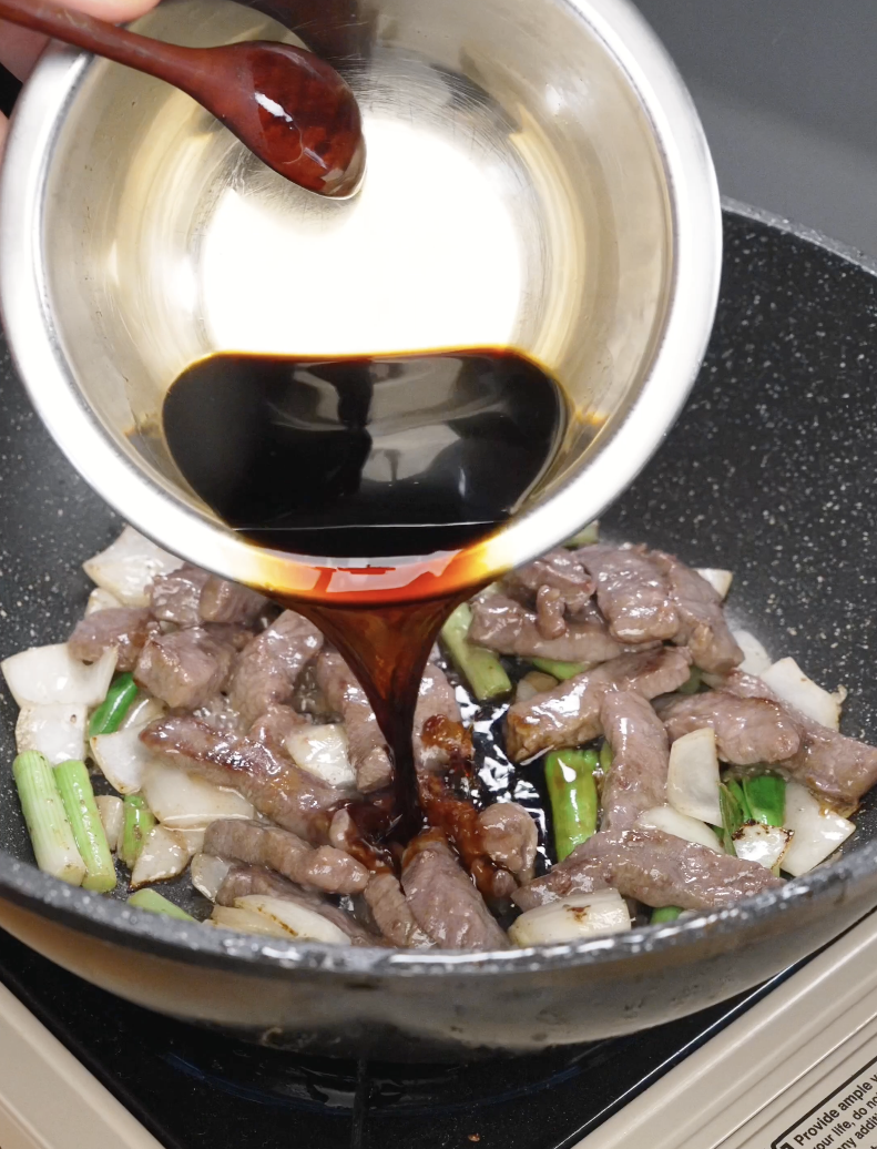 Thick soy-based sauce being poured over seared beef, onions, and scallions in a pan, coating everything in the Mongolian Beef Recipe.