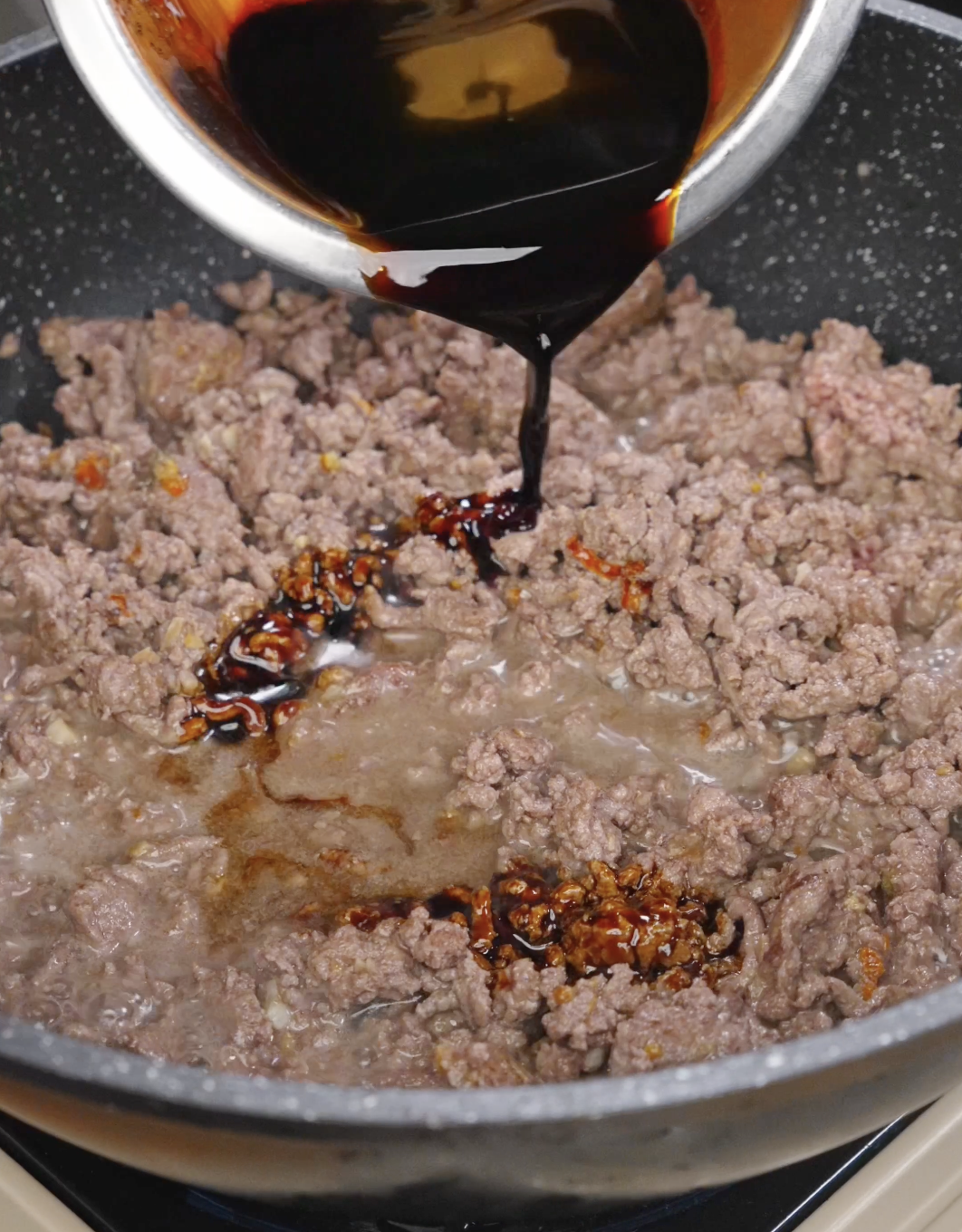 Prepared soy-based sauce being poured into browned ground beef, beginning the process of reducing and coating the meat.