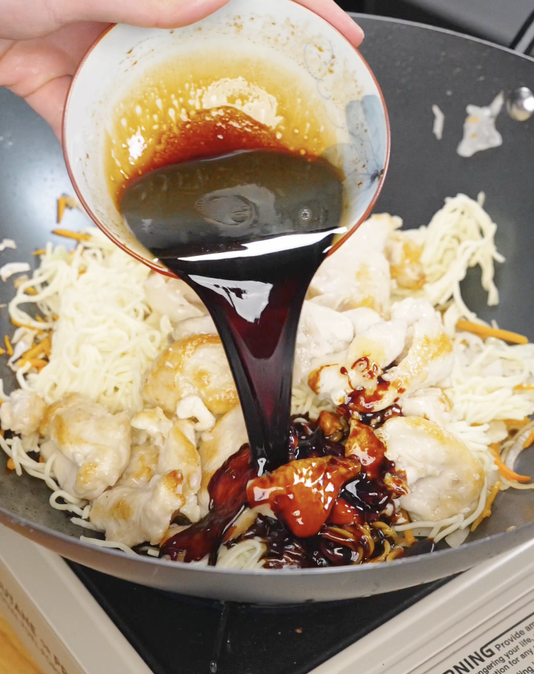 Thick soy-based sauce being poured over noodles and chicken in a pan, coating the chow mein with flavor.