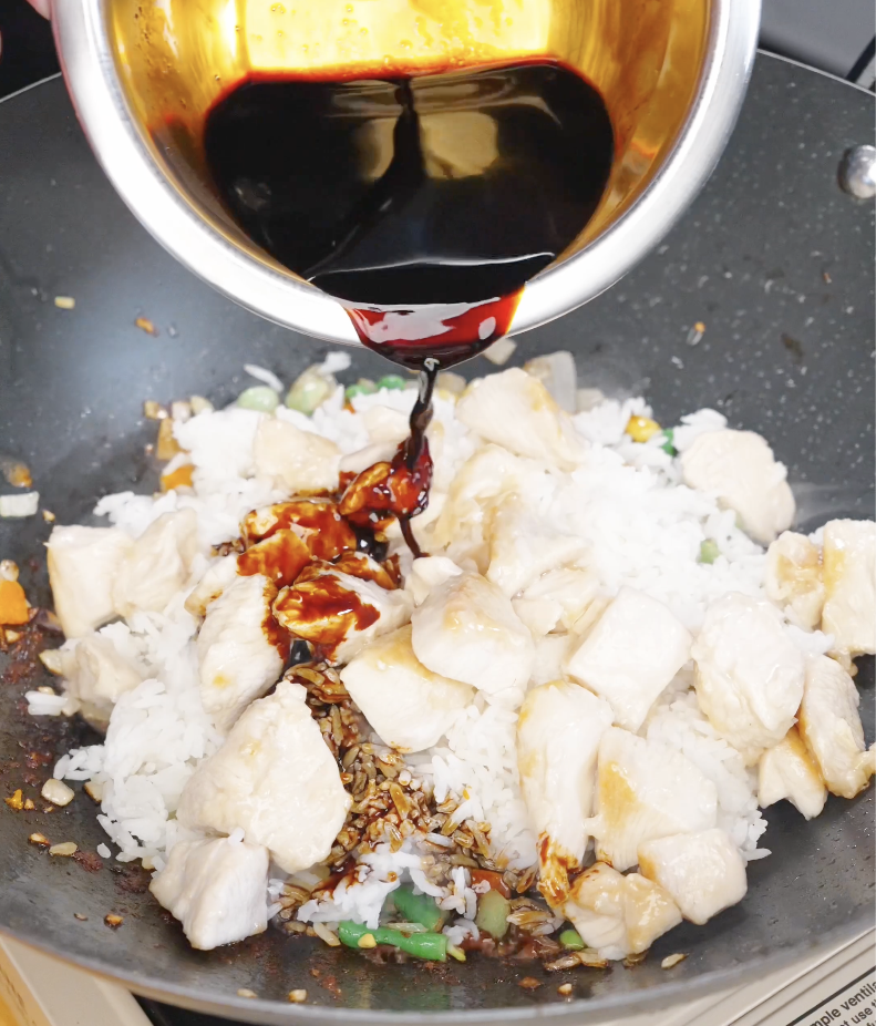 Soy-based sauce being poured over rice and cooked chicken in a pan, coating the fried rice with flavor.