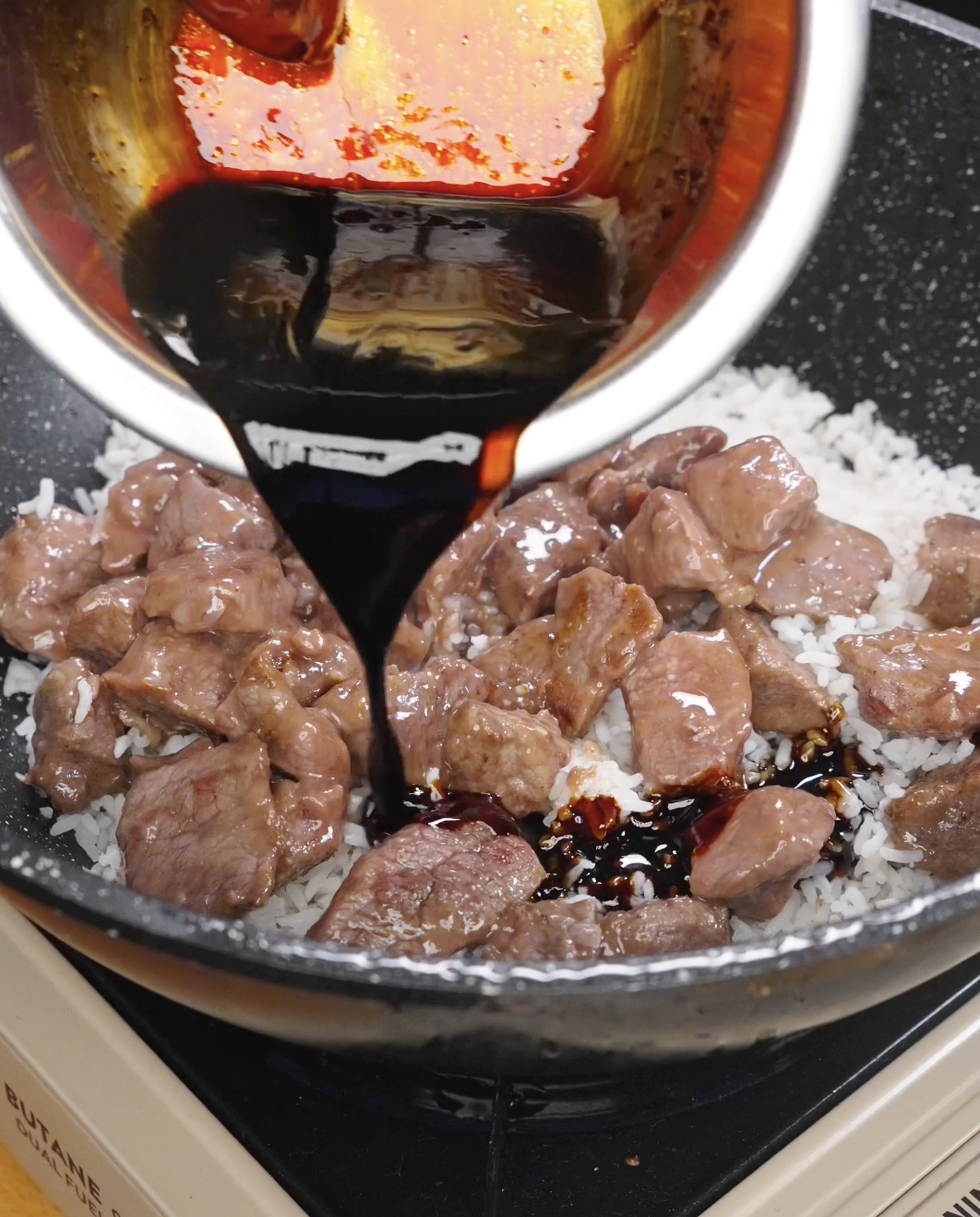 Dark soy-based sauce being poured over seared beef and day-old rice in a pan, coating the ingredients to create rich garlic beef fried rice.