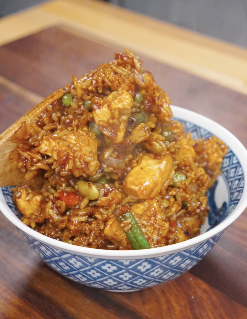 Wooden spoon scooping and serving chicken fried rice into a bowl, showing glossy rice with tender chicken and mixed vegetables coated in sauce.