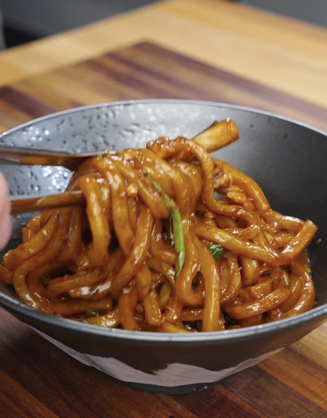 Freshly cooked garlic chicken noodles being lifted and served into a bowl, coated in a glossy soy-based sauce with tender chicken and scallions, highlighting the thick udon texture and rich savory finish.
