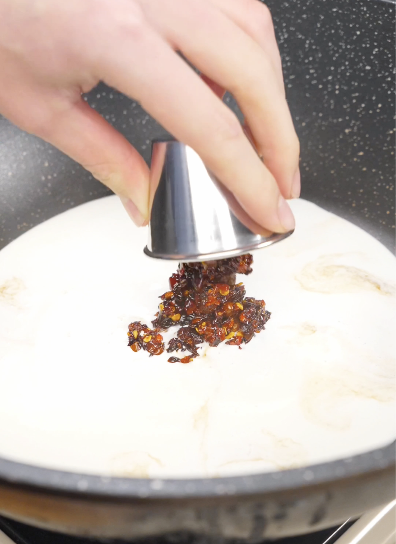 Chili oil being added to a skillet with heavy cream and seasonings to create the rich spicy sauce for Creamy Chili Oil Noodles Recipe.