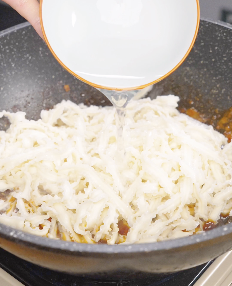 Reserved noodle water being poured into a skillet filled with cooked noodles and gochujang sauce. The starchy water helps emulsify the sauce and creates a silky texture that clings to the noodles.