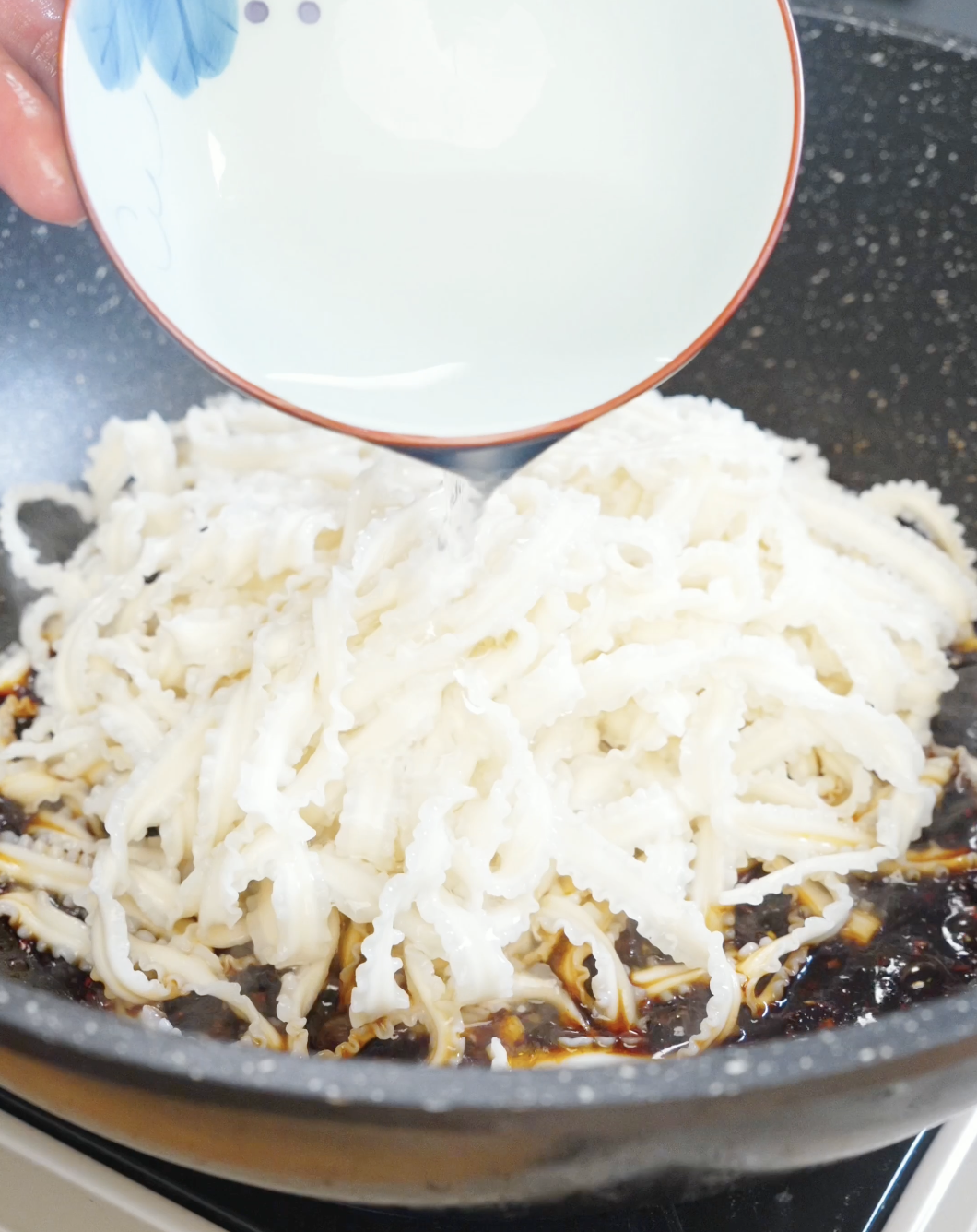 Reserved noodle water being poured into spicy garlic noodles in a skillet to help emulsify the sauce and coat every strand evenly.