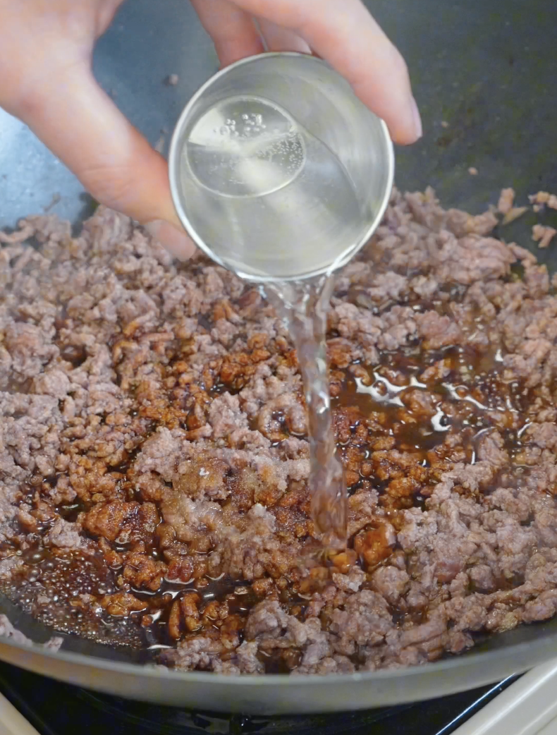 Sake being poured into seasoned ground beef to help create a glossy sauce in the Japanese Ground Beef Egg Bowl Recipe.