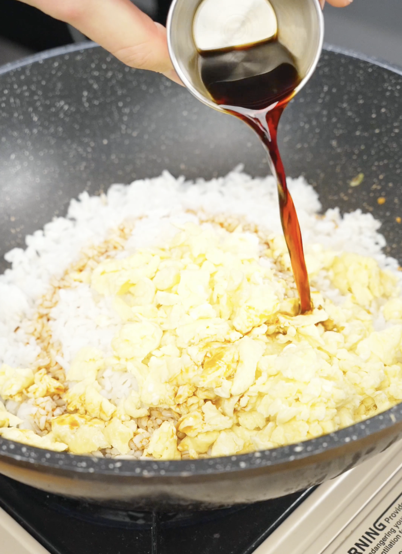 Soy sauce being poured over rice and scrambled eggs in a skillet while making Egg Fried Rice. The soy sauce seasons the rice and gives the dish its signature savory flavor and golden color.