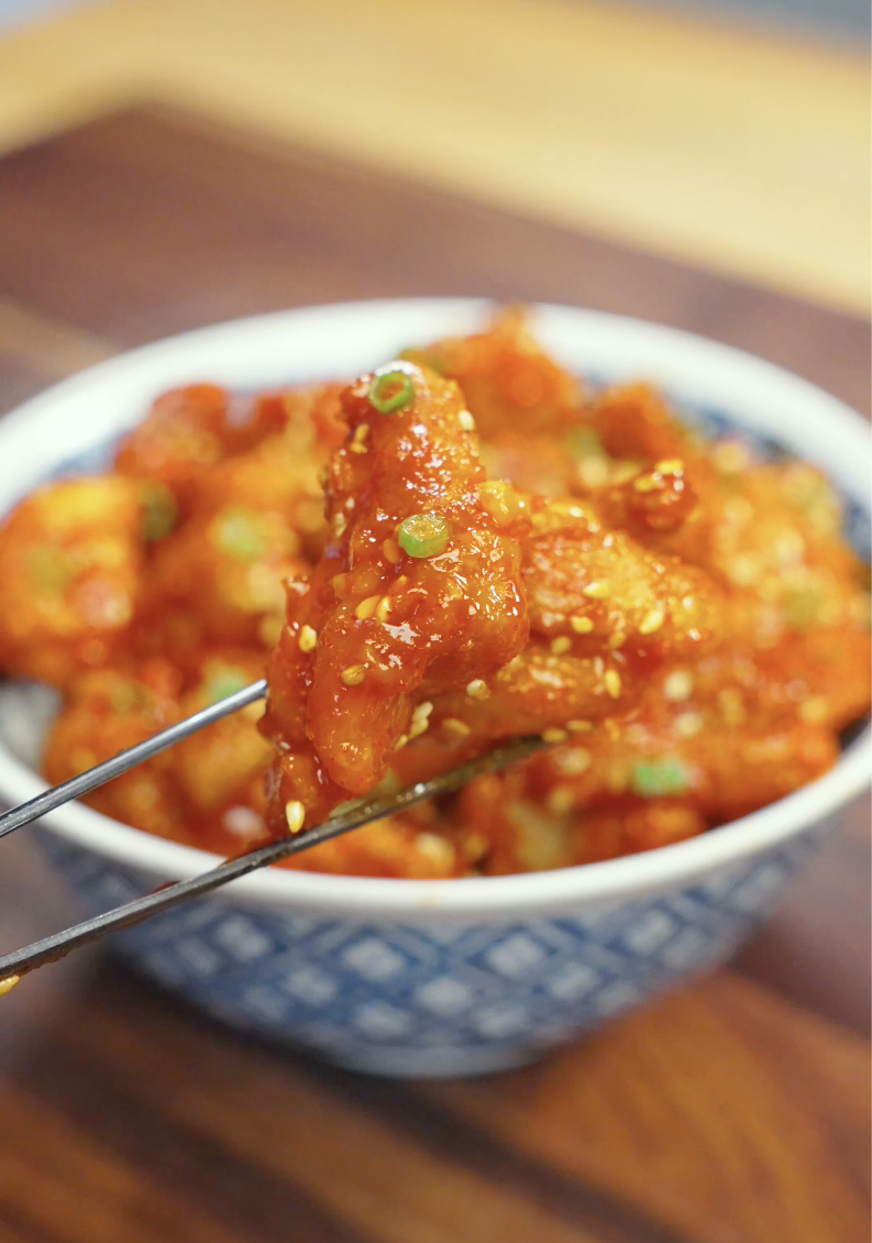 Close-up of a piece of Crispy Gochujang Chicken held with chopsticks, showcasing the thick sticky gochujang glaze, crunchy coating, and sesame seed garnish. Perfect for an easy Korean-inspired weeknight dinner.