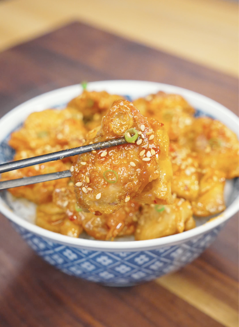Close-up of a crispy piece of Hot Honey Chicken held with chopsticks above a bowl of rice. The chicken is coated in a glossy sweet and spicy honey glaze and garnished with sesame seeds and scallions for extra flavor.