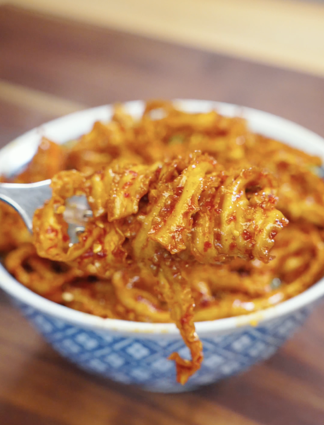 Fork lifting spicy garlic noodles coated in a glossy chili garlic sauce with visible chili flakes and sesame seeds.
