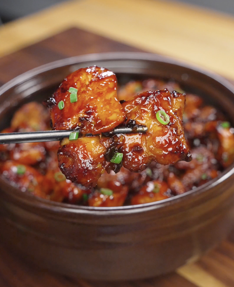 Close-up of a piece of Sticky Sweet Chicken held with chopsticks, showcasing the glossy sweet and savory glaze coating tender chicken thigh pieces. The sticky sauce creates the perfect balance of richness and sweetness in every bite.