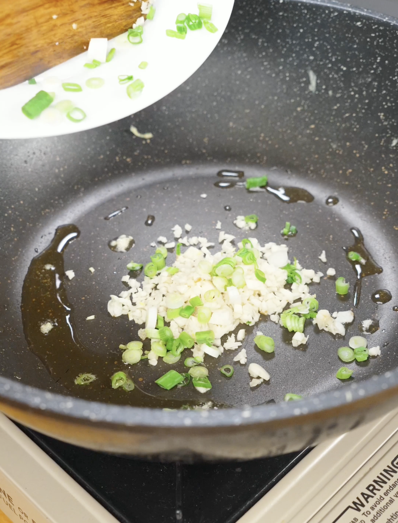 Minced garlic and sliced scallion whites cooking in a skillet with oil to create the flavor base for Egg Fried Rice. Saut&eacute;ing these aromatics first infuses the oil and enhances the overall flavor of the dish.