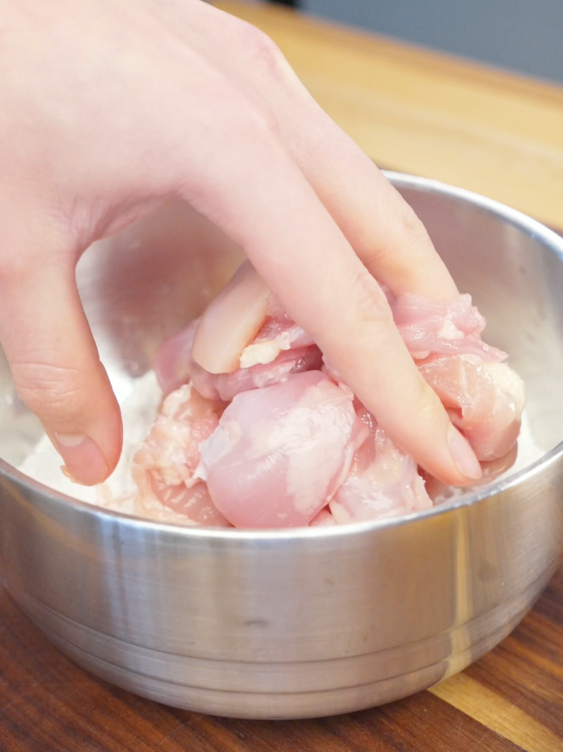 Cubed chicken thighs being added to a bowl of seasoned flour and cornstarch coating before air frying. Thoroughly coating the chicken helps create a crispy exterior without deep frying.