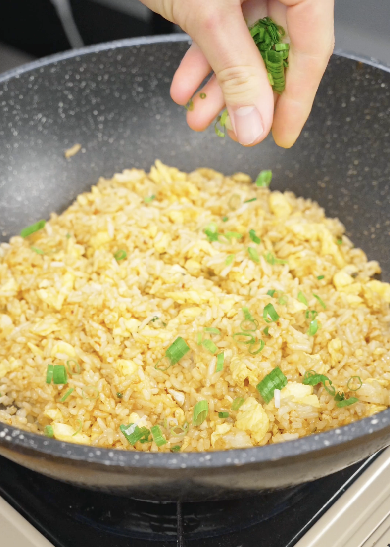 Fresh scallion greens being sprinkled into a skillet of Egg Fried Rice during the final stage of cooking. Adding the greens at the end preserves their freshness and bright flavor.