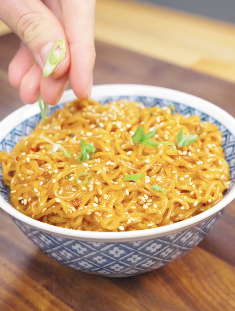 Fresh scallions being sprinkled over a bowl of Creamy Chili Oil Noodles Recipe finished with sesame seeds and a rich spicy cream sauce.