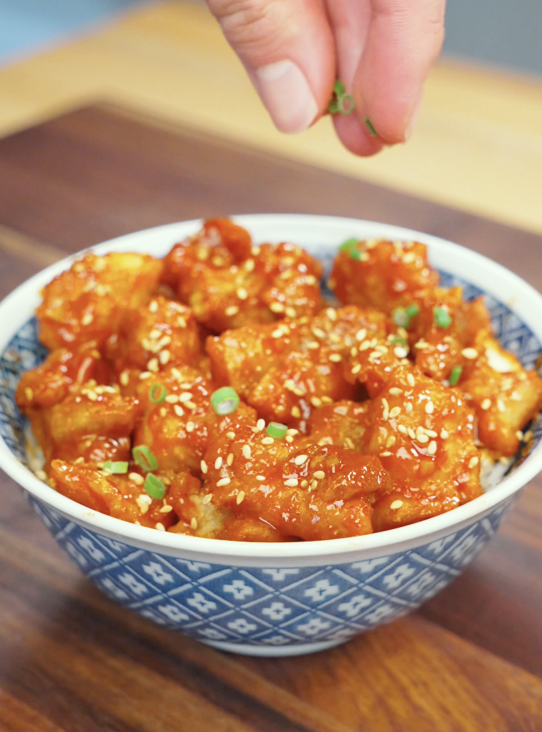 Fresh scallions being sprinkled over a bowl of Crispy Gochujang Chicken finished with sesame seeds and sticky gochujang glaze. This final garnish adds freshness, color, and texture to the dish.