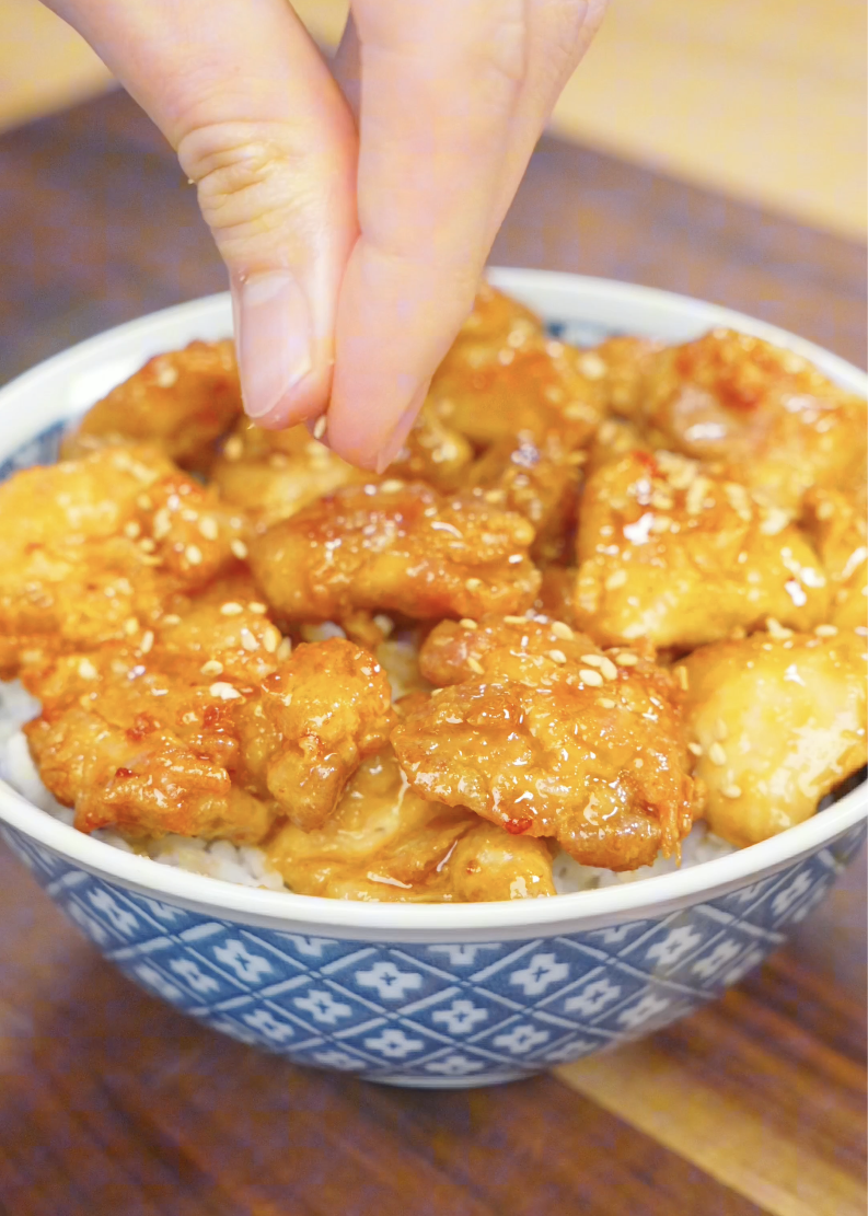 Sesame seeds being sprinkled over Crispy Hot Honey Chicken served on a bowl of rice. The finishing garnish adds texture, flavor, and visual appeal to this easy air fryer chicken dinner.