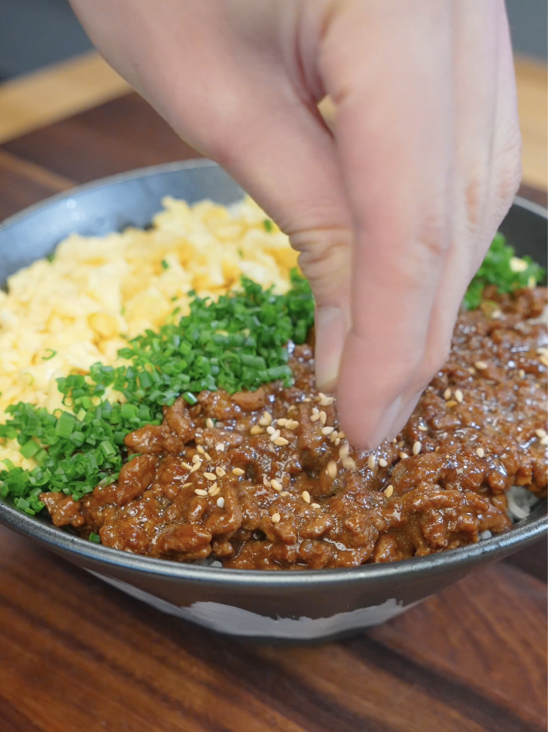 Sesame seeds being sprinkled over glazed ground beef, scrambled eggs, and chives in the finished Japanese Ground Beef Egg Bowl Recipe.