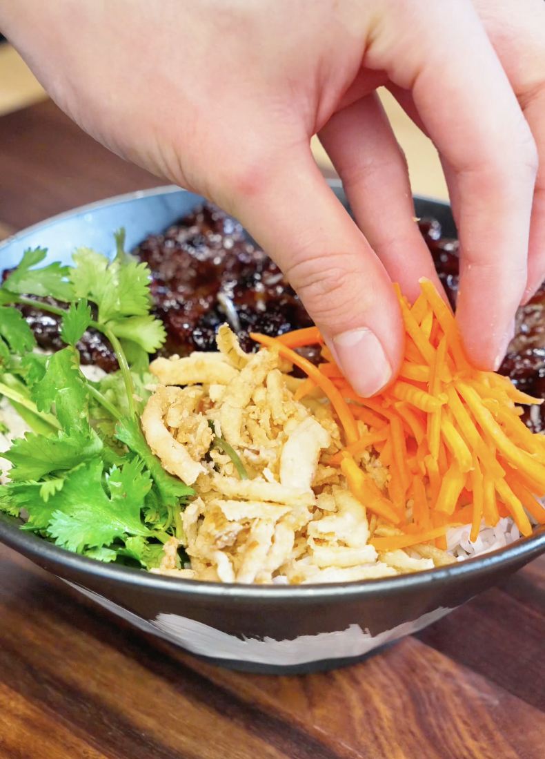 Fresh shredded carrots being added to a lemongrass beef rice bowl with cilantro and crispy onions for extra texture and vibrant flavor.