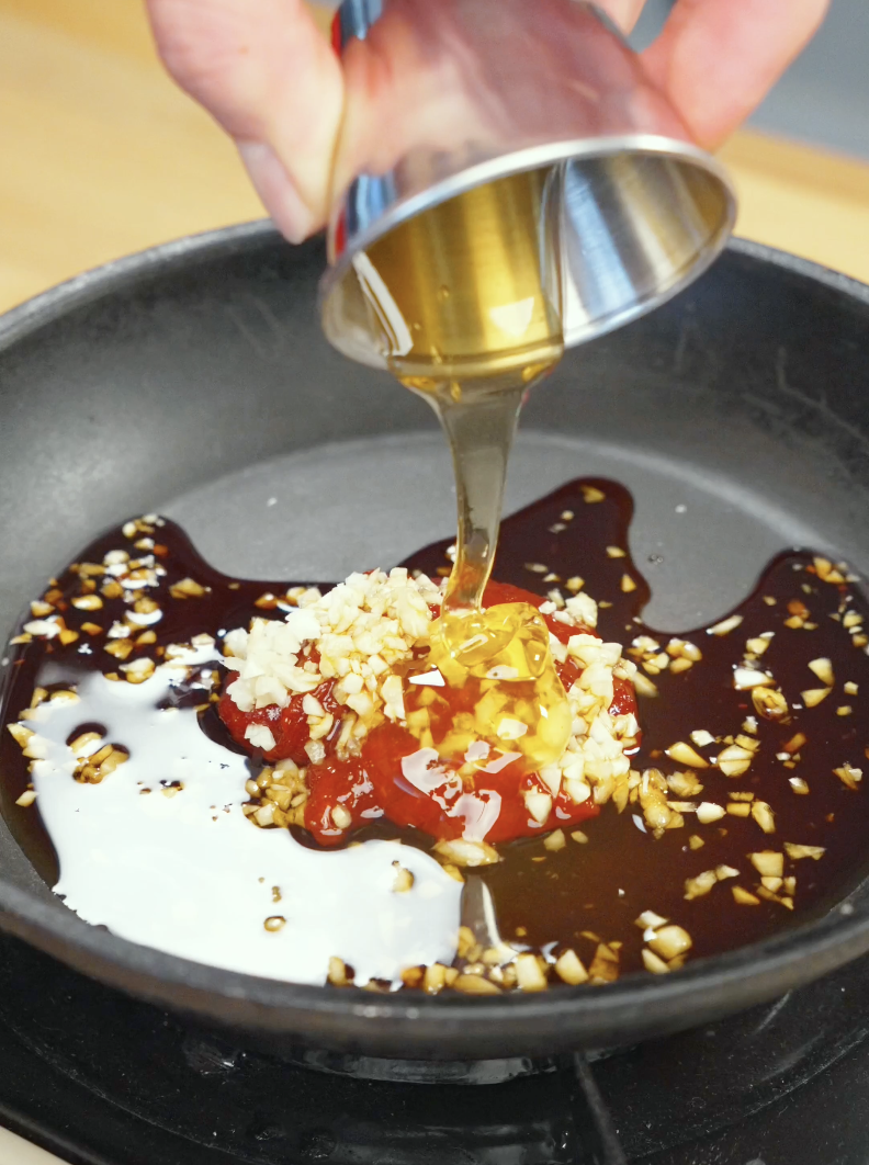 Honey being poured into a skillet with gochujang, garlic, soy sauce, rice vinegar, and sesame oil to create a sweet, spicy, and savory glaze for Crispy Gochujang Chicken Recipe.