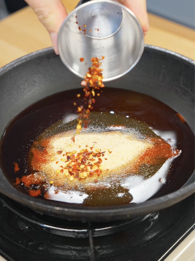 Chili flakes being added to a skillet containing honey, soy sauce, rice vinegar, sriracha, and garlic powder. Simmering these ingredients together creates a sticky sweet and spicy hot honey glaze for the chicken.