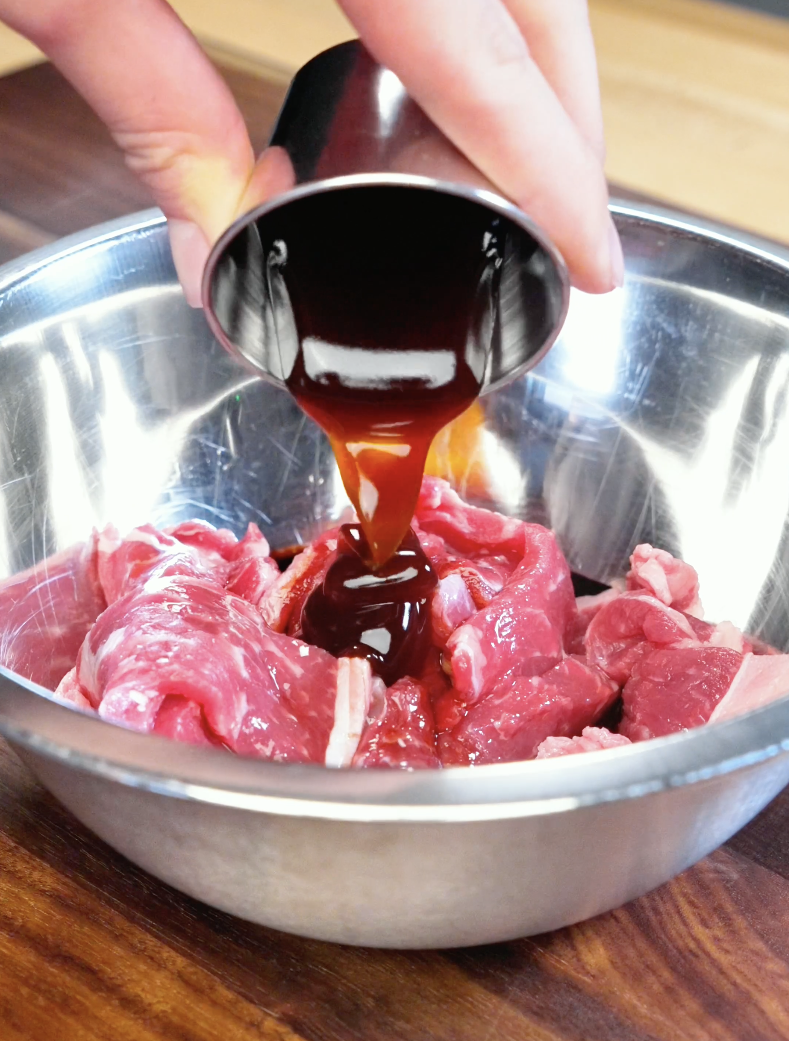 Dark soy sauce marinade being poured over thinly sliced strip steak with lemongrass and garlic to prepare flavorful Vietnamese-style Lemongrass Beef.