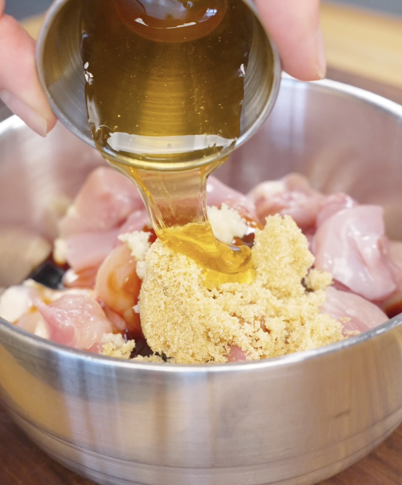 Honey being poured over cubed chicken thighs along with soy sauces, oyster sauce, garlic, brown sugar, rice vinegar, and sesame oil. This flavorful marinade forms the foundation of the Sticky Sweet Chicken Recipe and helps create its signature glaze.