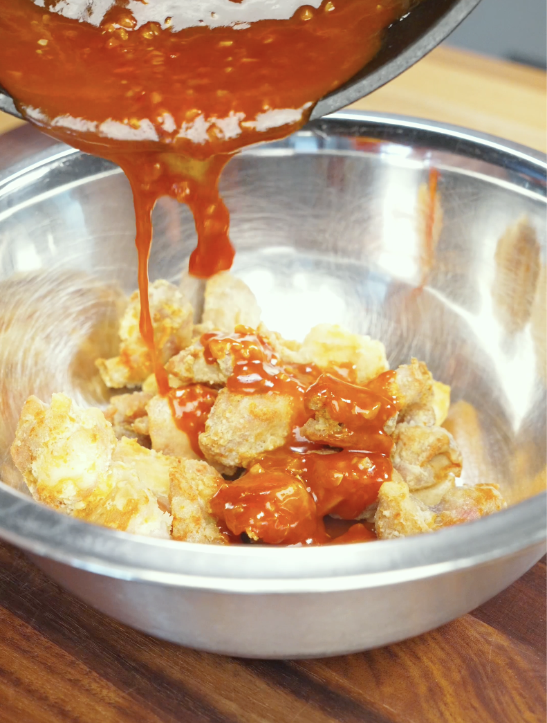Thick homemade gochujang sauce being poured over crispy air-fried chicken pieces in a mixing bowl. The glossy Korean-inspired glaze coats every piece for maximum flavor.