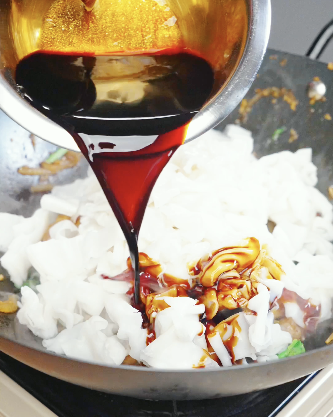 Savory Pad Kee Mao sauce being poured over wide rice noodles in a wok for a Drunken Noodles Recipe. The noodles absorb the sauce and become glossy and flavorful.